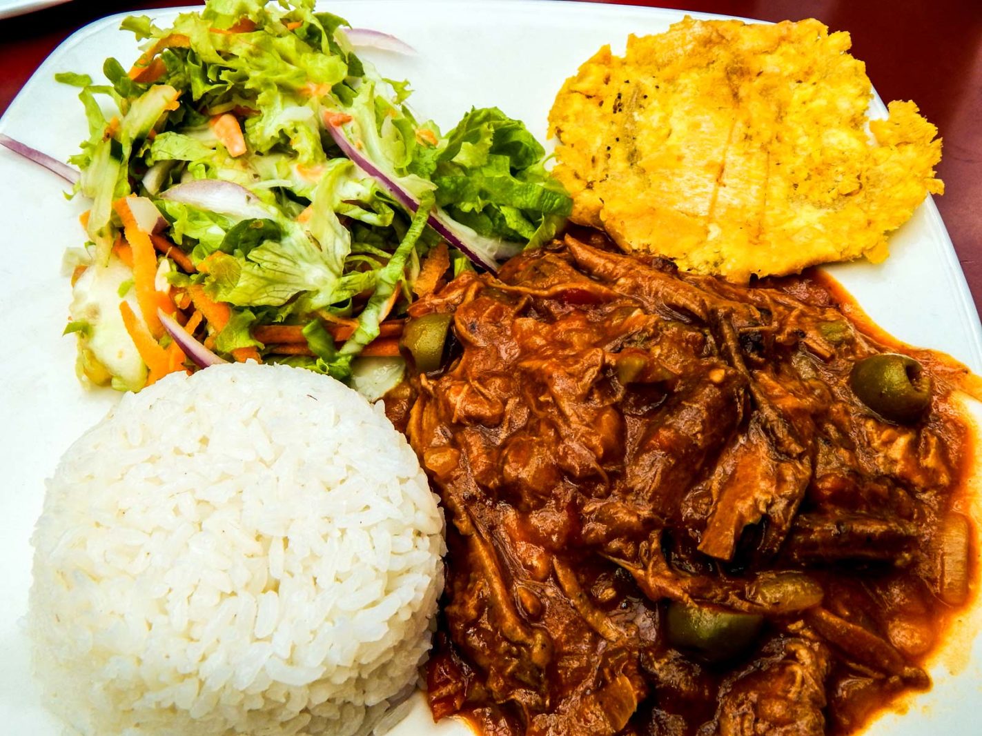 A white plate of Cuban Ropa Vieja, showing tender shredded beef in a vibrant tomato and pepper sauce, next to a side of white rice.