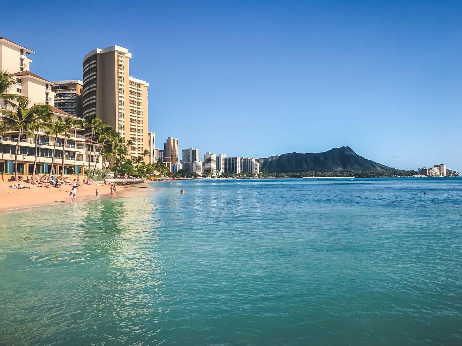 A sunny day on Waikiki Beach in Oahu, with surfers in the turquoise water and the iconic Diamond Head crater in the background.