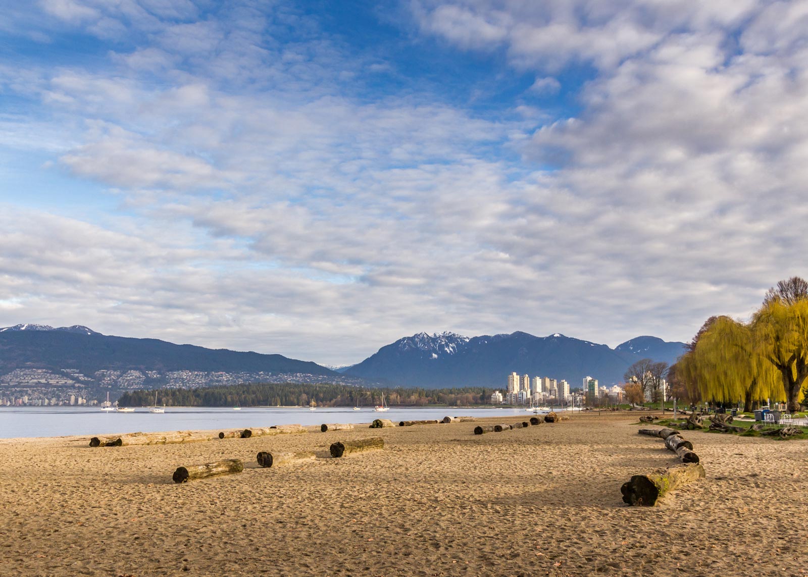 A wide, sunny view of Kitsilano Beach in Vancouver, with the downtown skyline visible across the water.