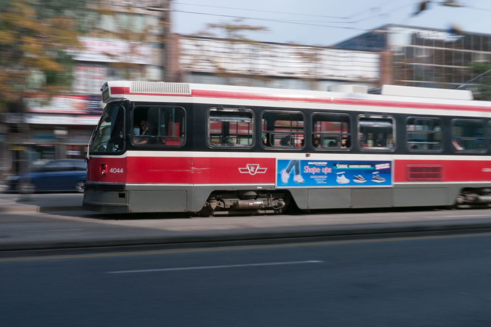 A classic red TTC streetcar driving down a busy Queen Street West in Toronto, with shops and people lining the sidewalks.